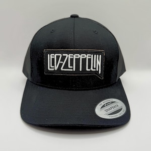 May include: Black snapback hat with a mesh back and a curved brim. The front features a rectangular patch with the band name "LED ZEPPELIN" in white lettering. A silver "SNAPBACK" sticker is on the brim.