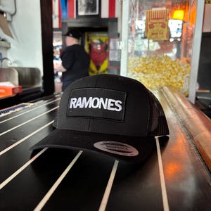 K&ouml;nnte beinhalten: Schwarze Trucker-M&uuml;tze mit Mesh-R&uuml;ckseite und gebogenem Schirm. Die M&uuml;tze hat einen rechteckigen Aufn&auml;her mit dem Wort "RAMONES" in wei&szlig;en Blockbuchstaben. Ein Snapback-Aufkleber ist auf dem Schirm sichtbar.