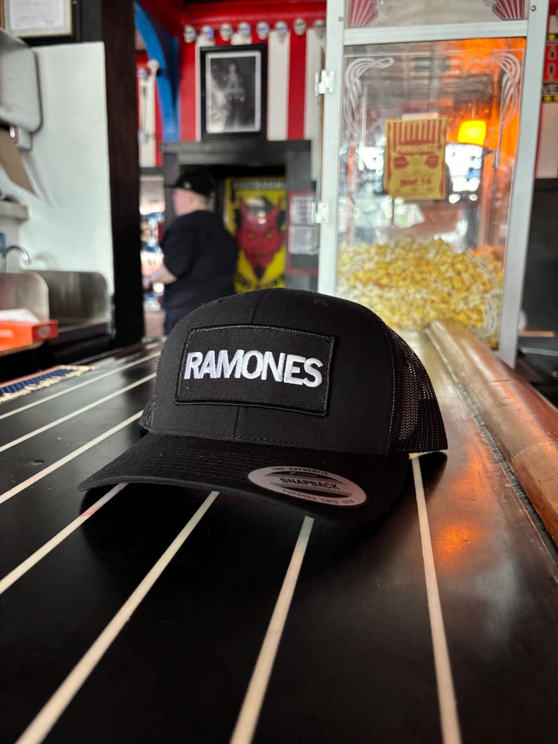 K&ouml;nnte beinhalten: Schwarze Trucker-M&uuml;tze mit Mesh-R&uuml;ckseite und gebogenem Schirm. Die M&uuml;tze hat einen rechteckigen Aufn&auml;her mit dem Wort "RAMONES" in wei&szlig;en Blockbuchstaben. Ein Snapback-Aufkleber ist auf dem Schirm sichtbar.