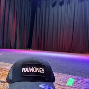 K&ouml;nnte beinhalten: Schwarze Trucker-M&uuml;tze mit dem Wort "RAMONES" in wei&szlig;en Buchstaben auf einem rechteckigen Aufn&auml;her. Die M&uuml;tze hat einen gebogenen Schirm und eine Mesh-R&uuml;ckseite. Die M&uuml;tze liegt auf einer Holzoberfl&auml;che.