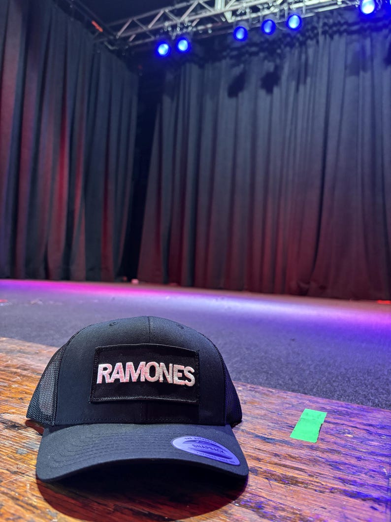 K&ouml;nnte beinhalten: Schwarze Trucker-M&uuml;tze mit dem Wort "RAMONES" in wei&szlig;en Buchstaben auf einem rechteckigen Aufn&auml;her. Die M&uuml;tze hat einen gebogenen Schirm und eine Mesh-R&uuml;ckseite. Die M&uuml;tze liegt auf einer Holzoberfl&auml;che.