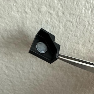 May include: A small, black, angular 3D-printed component featuring a circular, translucent, light-colored inlay at its center. The object is held by a pair of silver-toned metal tweezers, showcasing its textured surface and geometric shape.