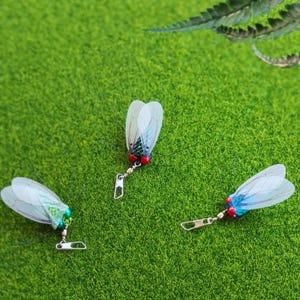 Peut inclure: Trois leurres de pêche artificiels en forme d'insectes, avec des ailes translucides et des accents rouges et verts. Chaque leurre est équipé d'un clip pivotant argenté pour l'attache à la ligne. Les leurres sont disposés sur une surface verte et herbeuse.