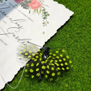 May include: A decorative butterfly with black and neon green spotted wings rests on a white card with floral accents and the text "You make my heart smile." The butterfly is set against a green, grassy background.