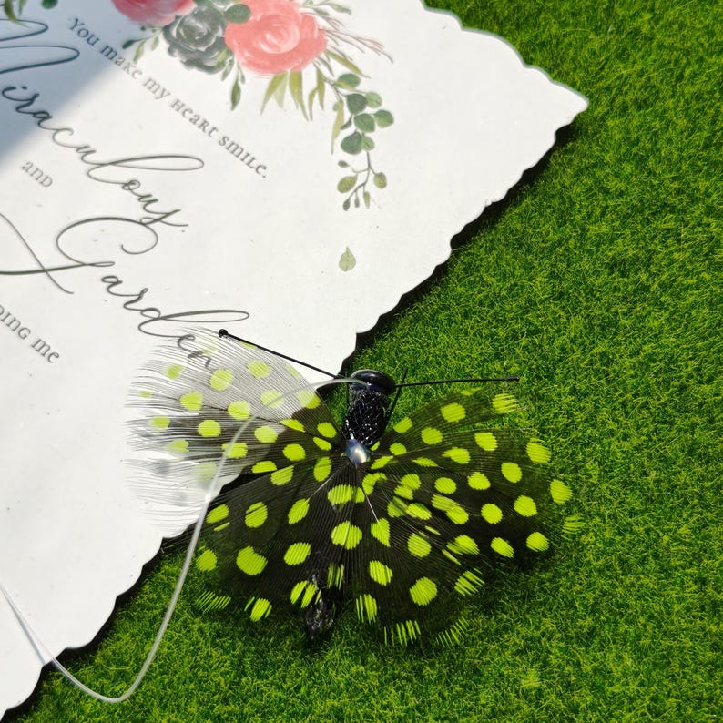 May include: A decorative butterfly with black and neon green spotted wings rests on a white card with floral accents and the text "You make my heart smile." The butterfly is set against a green, grassy background.