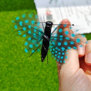 May include: A decorative butterfly with black body and wings made of feathers. The wings are white with turquoise polka dots. The butterfly is held by a person's fingers against a green background.