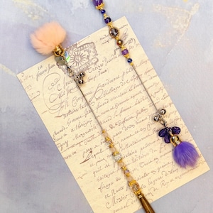 May include: Two decorative hair sticks with wooden handles and ornate beadwork. One features a pink fur accent, the other a purple fur accent. Both have gold and silver accents and are set against a vintage document.