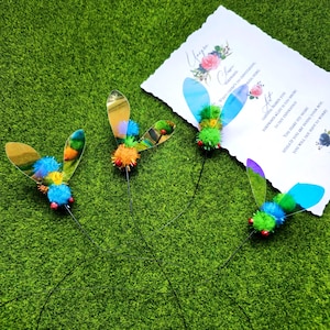 May include: Four decorative dragonfly ornaments with shimmering wings, vibrant pom-pom bodies, and slender wire stems. They are displayed on a green surface, alongside a card with floral designs and handwritten text.