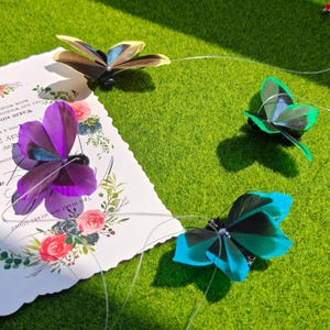 May include: Four colorful butterfly decorations with delicate wings are arranged on a green surface. One butterfly is purple, another is blue, a third is green, and the last is brown and yellow. A decorative card with floral designs and text is also present.