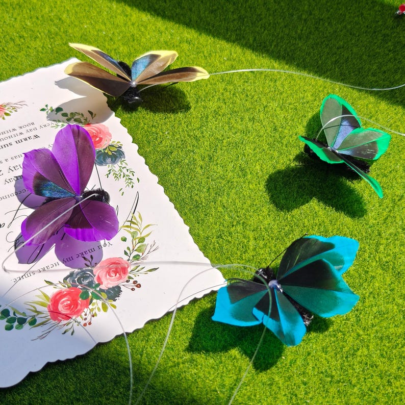 May include: Four colorful butterfly decorations with delicate wings are arranged on a green surface. One butterfly is purple, another is blue, a third is green, and the last is brown and yellow. A decorative card with floral designs and text is also present.