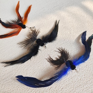 May include: Three decorative feather ornaments in shades of black, blue, and orange. Each ornament features a small, black velvet center with attached feathers and a silver-colored metal clip. The feathers are arranged to resemble birds in flight.