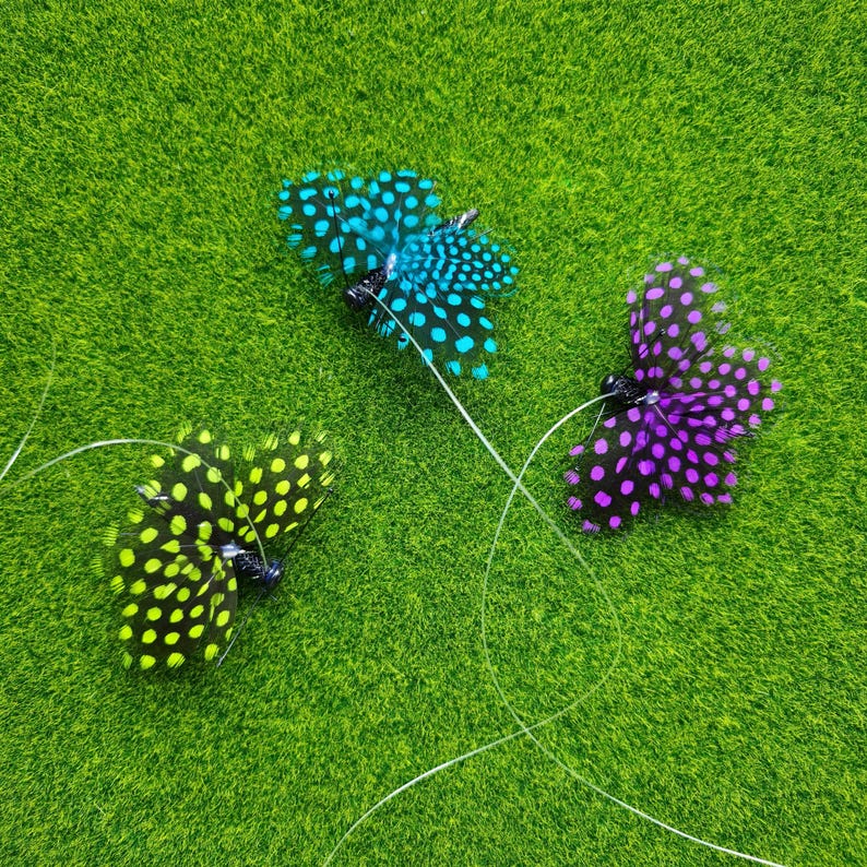 May include: Three feather butterflies in shades of blue, purple, and yellow with black spots. The butterflies are on a green surface, with thin, clear strings attached to them.