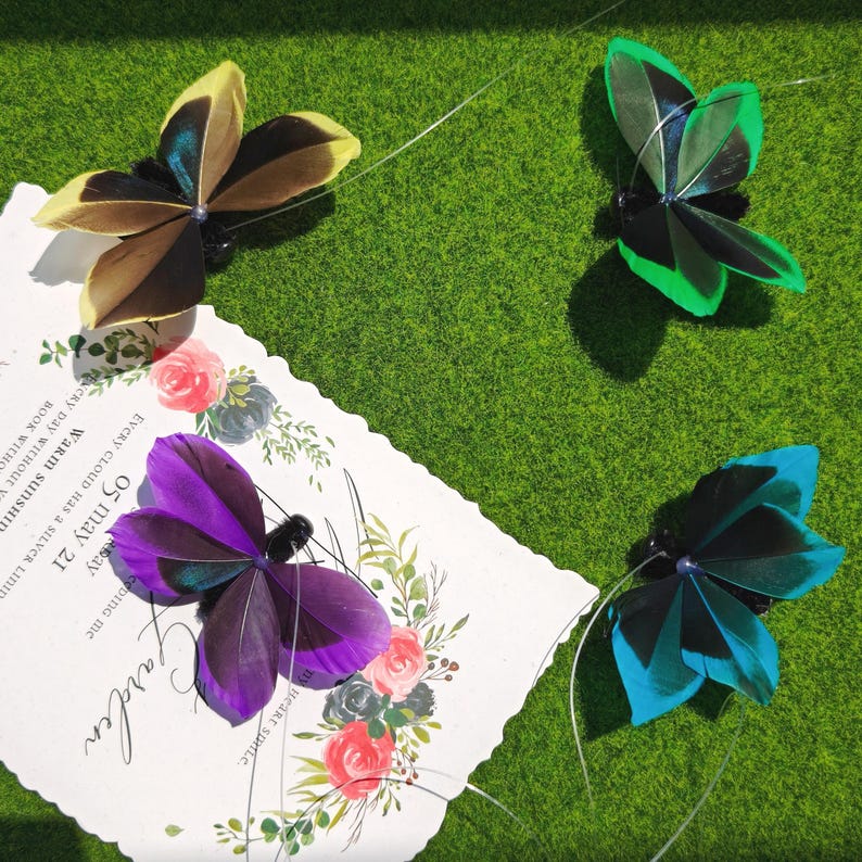 May include: Four decorative butterfly hair clips in yellow, green, blue, and purple. Each butterfly has a black center and delicate feather wings. The butterflies are arranged on a green surface with a floral card.