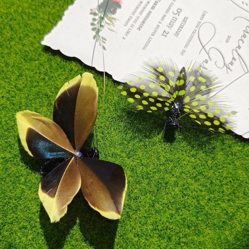 May include: Two decorative butterfly ornaments made of feathers. One butterfly has yellow, brown, and blue feathers. The other has black and white feathers with green polka dots. The butterflies are on a green surface with a greeting card.