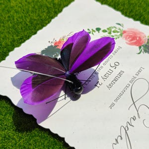 May include: A decorative butterfly with purple and black feathered wings rests on a white card. The card has floral accents and the text "05 may 21 Saturday". The butterfly has delicate antennae.