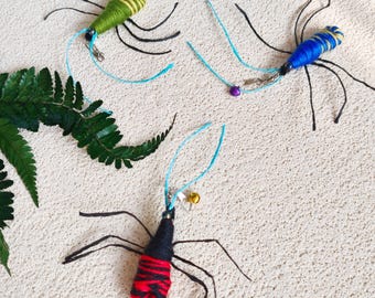 May include: Three handmade insect-shaped cat toys. One is green with yellow stripes, one is blue with yellow stripes, and the third is red and black striped. Each has black legs, a blue string, and a small bell or bead.