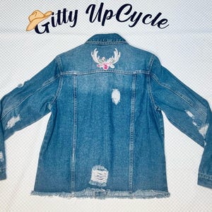 May include: A blue denim jacket with distressed details and a floral embroidered deer design on the back. The jacket has a frayed hem and is a great addition to any wardrobe.
