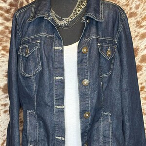 K&ouml;nnte beinhalten: Dunkelblaue Jeansjacke mit Kragen, Knopfleiste und mehreren Taschen. Die Jacke hat helle N&auml;hte und wird mit einem wei&szlig;en Tanktop und einer mehrschichtigen Silberkette getragen. Die Jacke wird auf einer schwarzen Schaufensterpuppe pr&auml;sentiert.