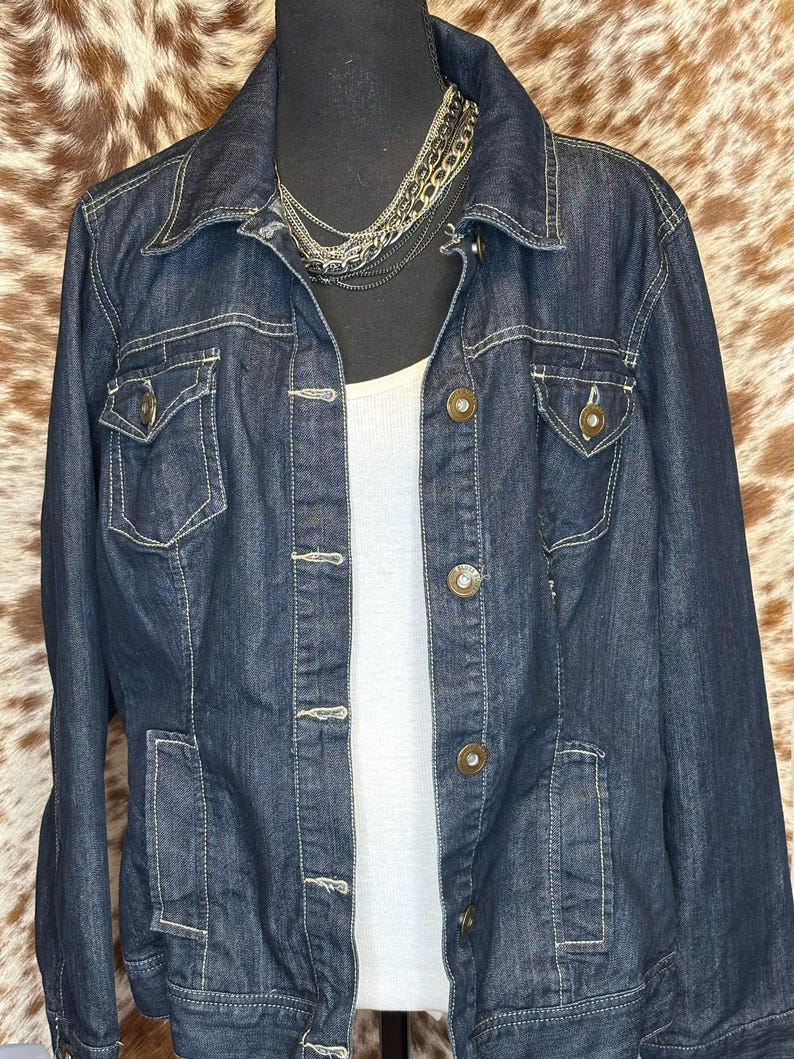 K&ouml;nnte beinhalten: Dunkelblaue Jeansjacke mit Kragen, Knopfleiste und mehreren Taschen. Die Jacke hat helle N&auml;hte und wird mit einem wei&szlig;en Tanktop und einer mehrschichtigen Silberkette getragen. Die Jacke wird auf einer schwarzen Schaufensterpuppe pr&auml;sentiert.