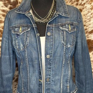 May include: A medium-wash denim jacket with a classic collar, button-down front, and two chest pockets. The jacket has a distressed look with frayed edges and yellow stitching details. A layered necklace is visible at the neckline.
