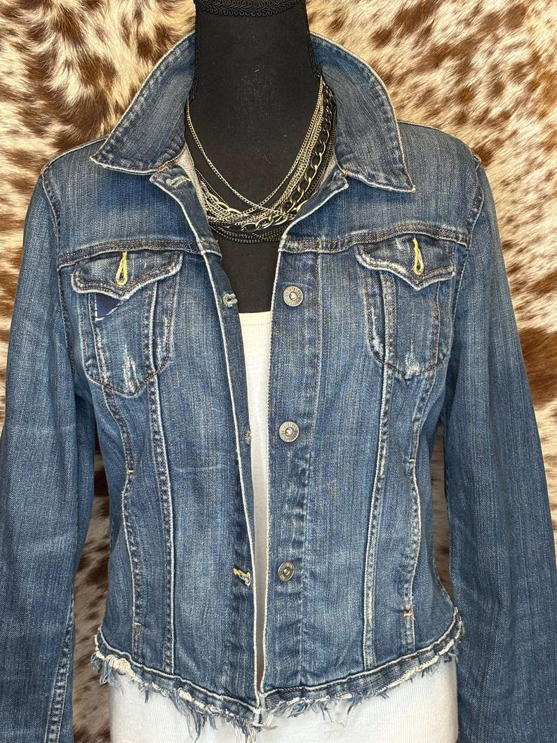 May include: A medium-wash denim jacket with a classic collar, button-down front, and two chest pockets. The jacket has a distressed look with frayed edges and yellow stitching details. A layered necklace is visible at the neckline.