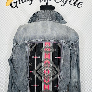 May include: A gray denim jacket with a black, pink, and gray patterned fabric patch on the back. The patch features a geometric design. The jacket has silver-toned buttons and pink embellishments.