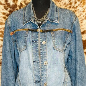 Puede incluir: Chaqueta vaquera azul claro con cuello cl&aacute;sico y cierre de botones. La chaqueta presenta dos bolsillos en el pecho y un ribete decorativo de flecos en tonos marr&oacute;n y negro. Se ve un collar de cadena plateada en capas.