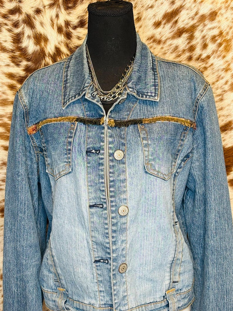 Puede incluir: Chaqueta vaquera azul claro con cuello cl&aacute;sico y cierre de botones. La chaqueta presenta dos bolsillos en el pecho y un ribete decorativo de flecos en tonos marr&oacute;n y negro. Se ve un collar de cadena plateada en capas.