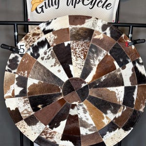 May include: A round rug with a patchwork design made from cowhide. The rug features a variety of brown, black, and white patches arranged in a radial pattern. The text "Gitty UpCycle" is visible in the background.