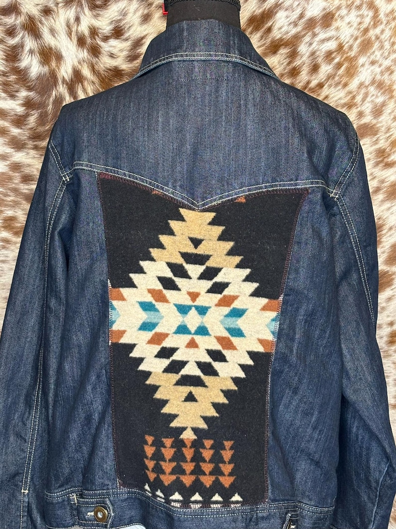 K&ouml;nnte beinhalten: Dunkelblaue Jeansjacke mit dekorativem R&uuml;ckenteil. Das Panel hat einen schwarzen Hintergrund und ein geometrisches Muster in Beige, T&uuml;rkis und Orange. Die Jacke hat wei&szlig;e N&auml;hte und einen klassischen Kragen.