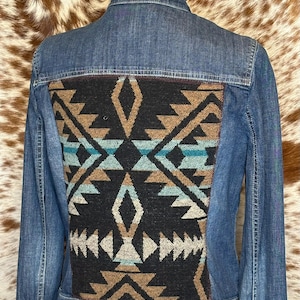 May include: Blue denim jacket with a decorative geometric pattern on the back. The pattern features shades of black, brown, and turquoise. The jacket has a classic collar and button closures.