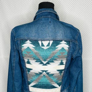 May include: A blue denim jacket with a geometric patterned back panel in teal, gray, and white. The jacket has a classic collar and button closures. The text "Gitty Up Cycle" is above the jacket.