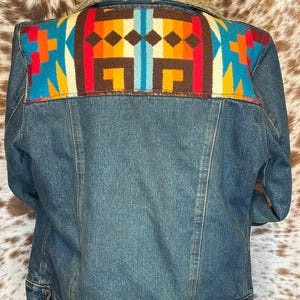 K&ouml;nnte beinhalten: Jeansjacke mit farbenfrohem, gemustertem Einsatz. Der Einsatz hat ein geometrisches Design in Blau-, Orange-, Rot- und Braunt&ouml;nen. Die Jacke hat einen Kragen aus beigem Kunstfell.