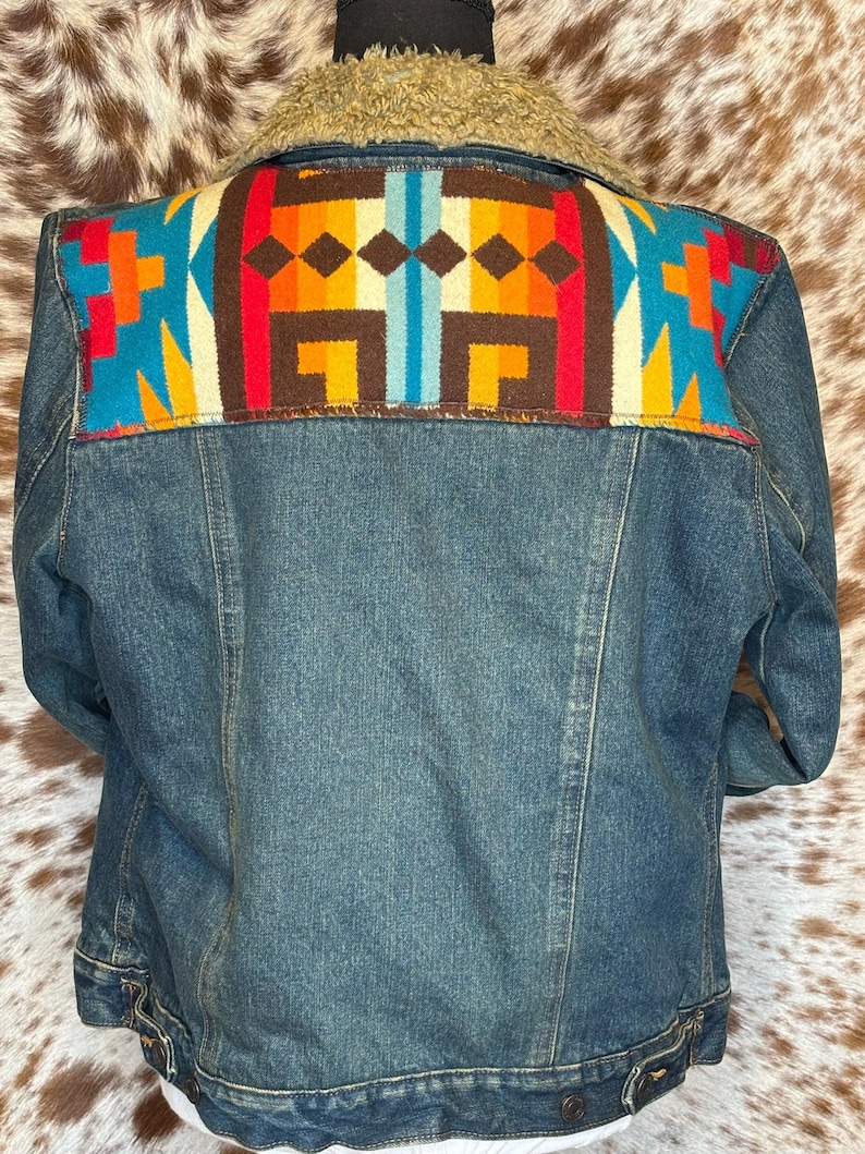 K&ouml;nnte beinhalten: Jeansjacke mit farbenfrohem, gemustertem Einsatz. Der Einsatz hat ein geometrisches Design in Blau-, Orange-, Rot- und Braunt&ouml;nen. Die Jacke hat einen Kragen aus beigem Kunstfell.