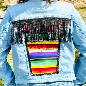 May include: A light blue denim jacket with a colorful striped fabric patch on the back. The patch is surrounded by a black fringe trim with rhinestones.