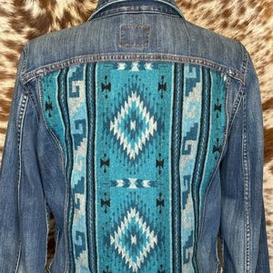 May include: A denim jacket with a turquoise and white patterned back panel. The back panel features a geometric design with diamond and stripe patterns. The jacket has a frayed hemline and a classic denim jacket design.