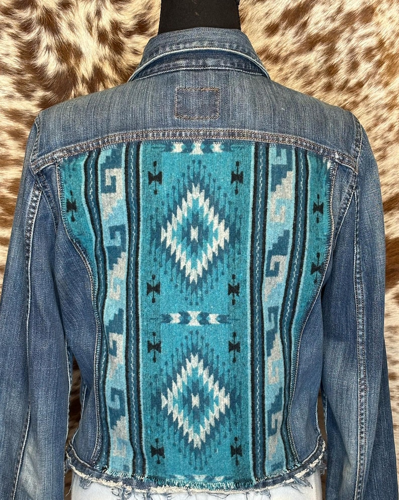 May include: A denim jacket with a turquoise and white patterned back panel. The back panel features a geometric design with diamond and stripe patterns. The jacket has a frayed hemline and a classic denim jacket design.