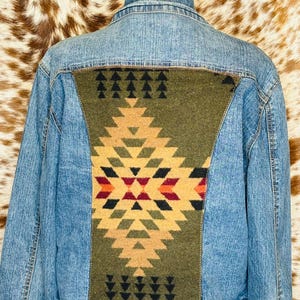 Puede incluir: Chaqueta vaquera azul claro con un panel decorativo verde oliva en la espalda con un patr&oacute;n geom&eacute;trico en tonos beige, rojo y negro. La chaqueta tiene un cintur&oacute;n de cuero marr&oacute;n.