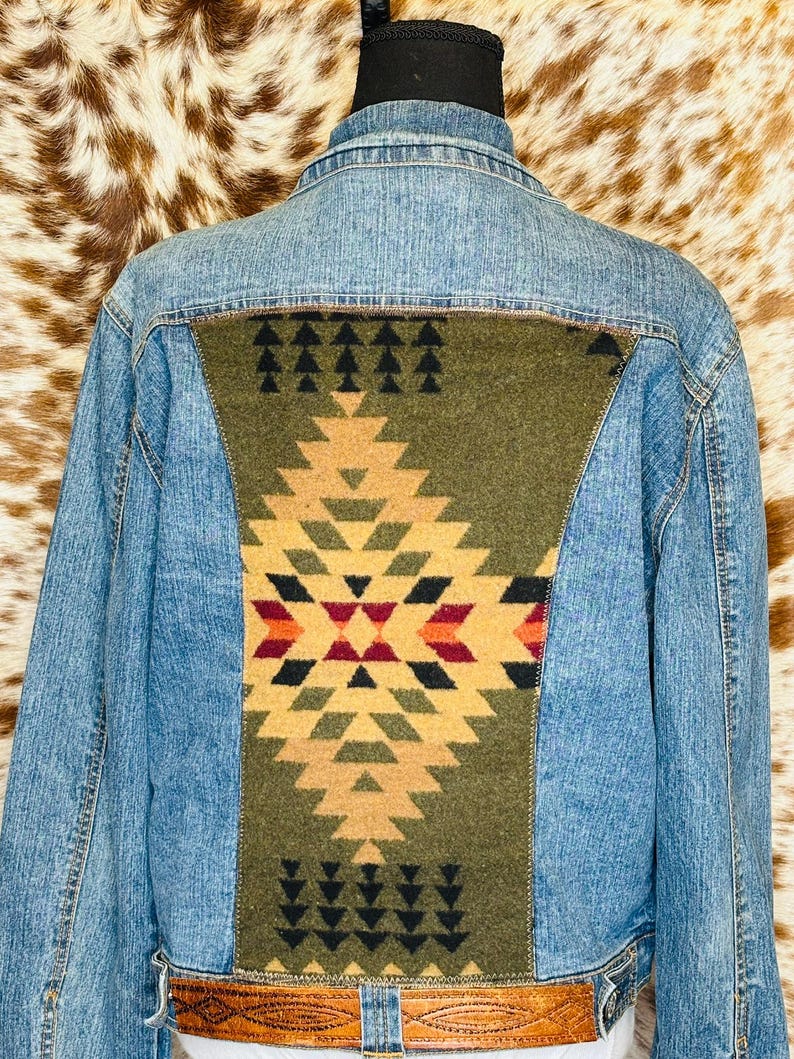 Puede incluir: Chaqueta vaquera azul claro con un panel decorativo verde oliva en la espalda con un patr&oacute;n geom&eacute;trico en tonos beige, rojo y negro. La chaqueta tiene un cintur&oacute;n de cuero marr&oacute;n.
