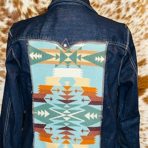 May include: Dark blue denim jacket with a colorful geometric design on the back. The design features shades of blue, orange, green, and cream. The jacket has a classic collar, button closures, and a tailored fit.