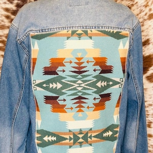 May include: Light blue denim jacket with a colorful geometric pattern on the back. The pattern features shades of teal, orange, brown, and cream. The jacket has a classic collar and button closures.