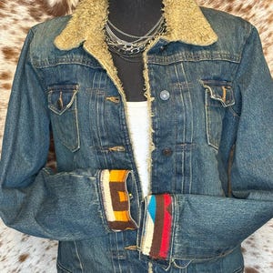K&ouml;nnte beinhalten: Blaue Jeansjacke mit beigem Kunstfellkragen und gemusterten Manschetten. Die Jacke hat zwei Brusttaschen und eine Knopfleiste. Die Manschetten zeigen geometrische Designs in Orange, Braun und Blau.