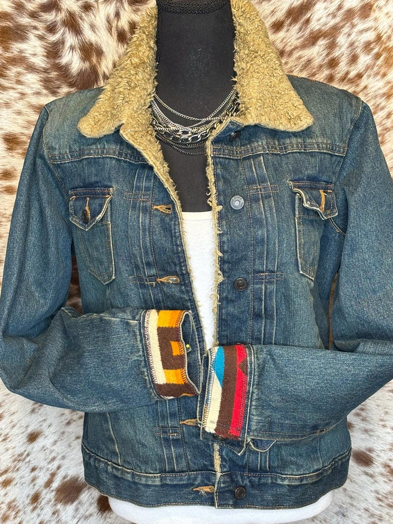 K&ouml;nnte beinhalten: Blaue Jeansjacke mit beigem Kunstfellkragen und gemusterten Manschetten. Die Jacke hat zwei Brusttaschen und eine Knopfleiste. Die Manschetten zeigen geometrische Designs in Orange, Braun und Blau.