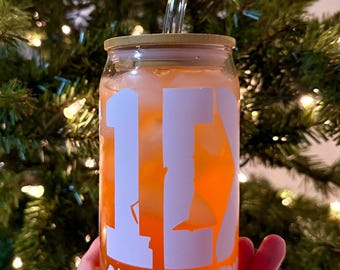 May include: A clear glass can with a gold lid and a white design that says "1D One Direction". The can is filled with a light orange liquid and has a clear glass straw.