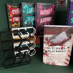 May include: A shelf displays three books from "The Rumious Love" trilogy, with titles like "Scythe & Sparrow" and "Butcher & Blackbird." A black and white book organizer sits in front. A book with a bloody cover is also visible.