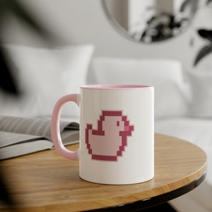 May include: A white ceramic mug with a pink rim and handle. The mug features a pink pixelated duck design.