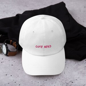 May include: White baseball cap with a pink embroidered phrase "CUTE NERD" on the front.