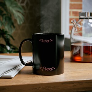 Op de afbeelding: Een zwarte keramische mok met de tekst "<tea>" en "/tea>" in roze op de voorkant. Stoom stijgt op uit de mok.