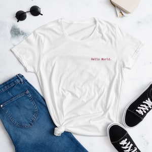 Op de afbeelding: Wit T-shirt met een roze "Hello World." tekst op de voorkant. Het T-shirt ligt op een wit oppervlak met een spijkerbroek, een zonnebril en zwarte sneakers.