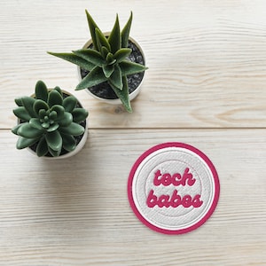 May include: A white round patch with a pink border and the text "tech babes" in pink lettering. The patch is on a light wood surface with two small potted succulents.